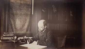 Henry Adams seated at desk in dark coat, writing Photograph