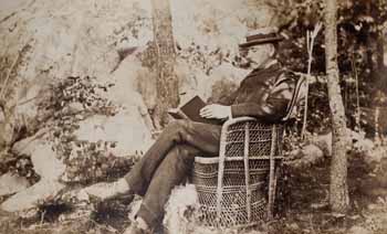 James Lowndes at Beverly Farms, seated outdoors in wicker chair, reading book, with dog at feet Photograph